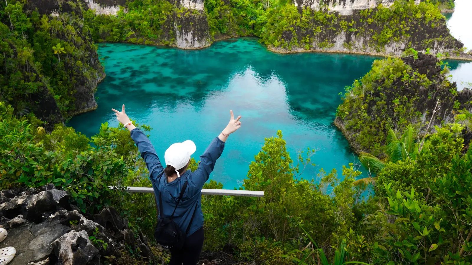 Raja Ampat – traveller with arms raised at a viewpoint above a turquoise karst lagoon – Indonesia